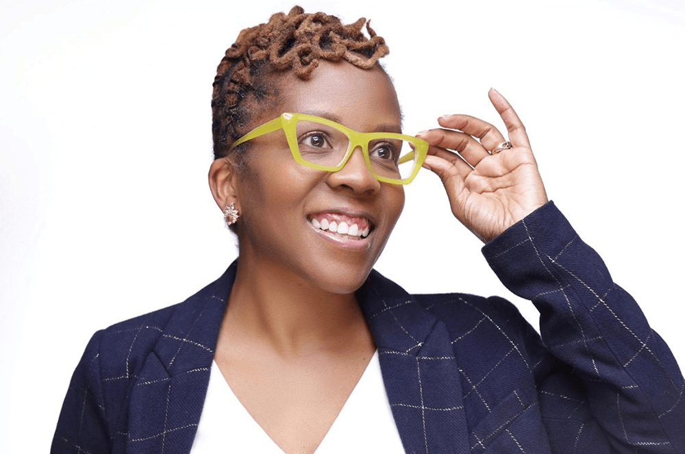 Woman in blue blazer reaching up to yellow glasses and smiling