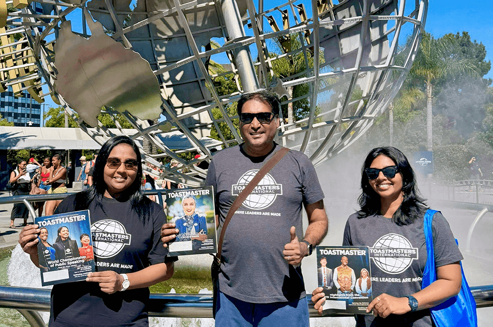 Two women and a man posing with the Toastmaster magazine in front of the Universal Studios Hollywood globe