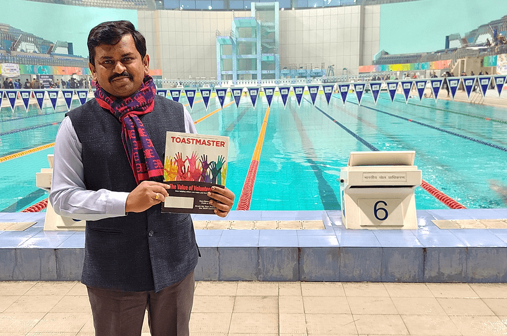 Man posing with Toastmaster magazine at indoor swimming pool