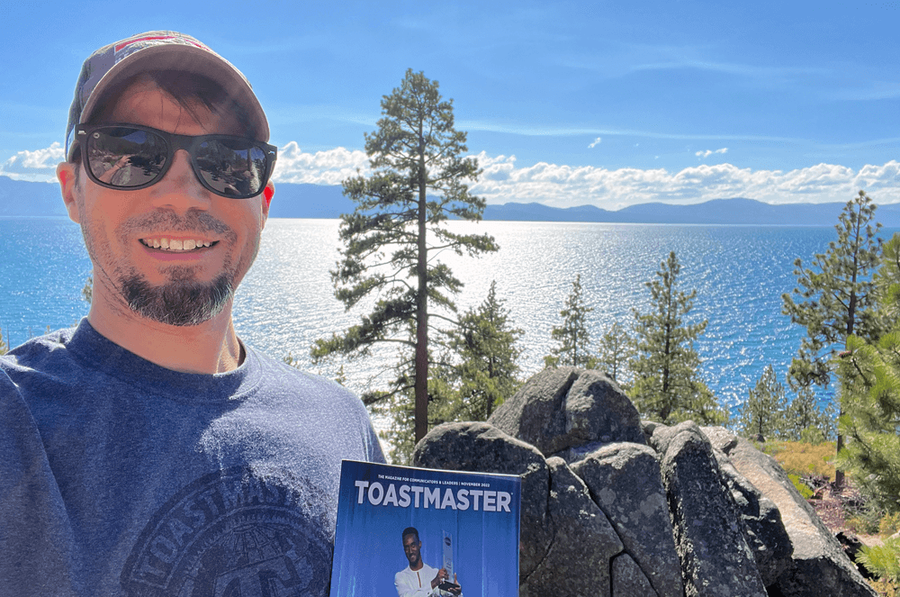 Man smiling and holding a magazine in front of a lake