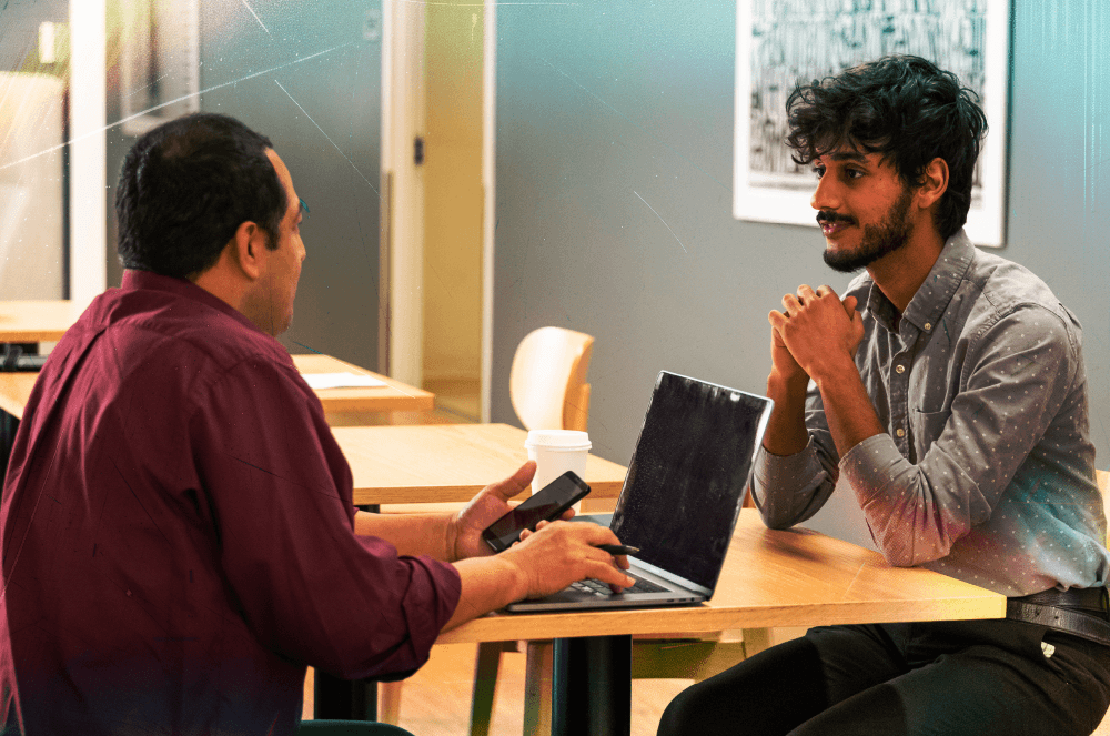 Two men sitting across from each other at a table with a phone and a laptop