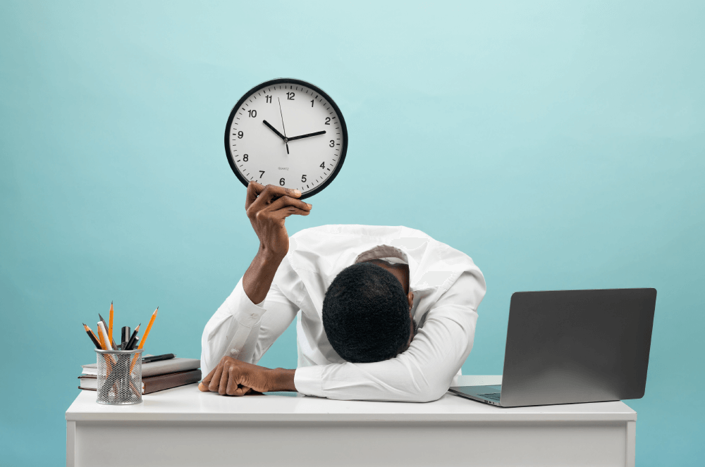A person holding a clock with their head on their desk