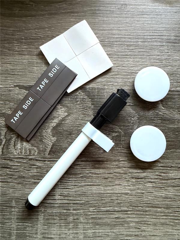 Magnetic and adhesive mounting strips, white magnets, and a black dry-erase pen scattered on a gray wood-grain background.