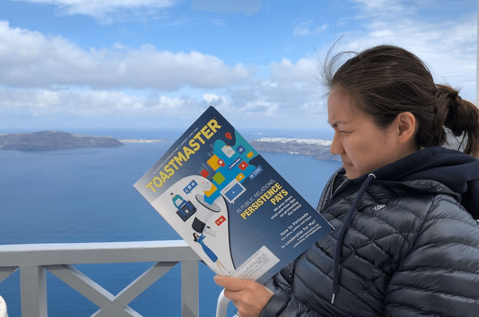 Lefan Zhuang of San Dimas, California, reads her Toastmaster magazine on the balcony of her hotel in Imerovigli, Greece. The white buildings in the background are part of the city of Oia.