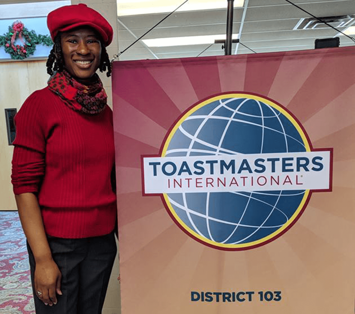 Cassandra lee in red shirt and hat standing next to Toastmasters banner