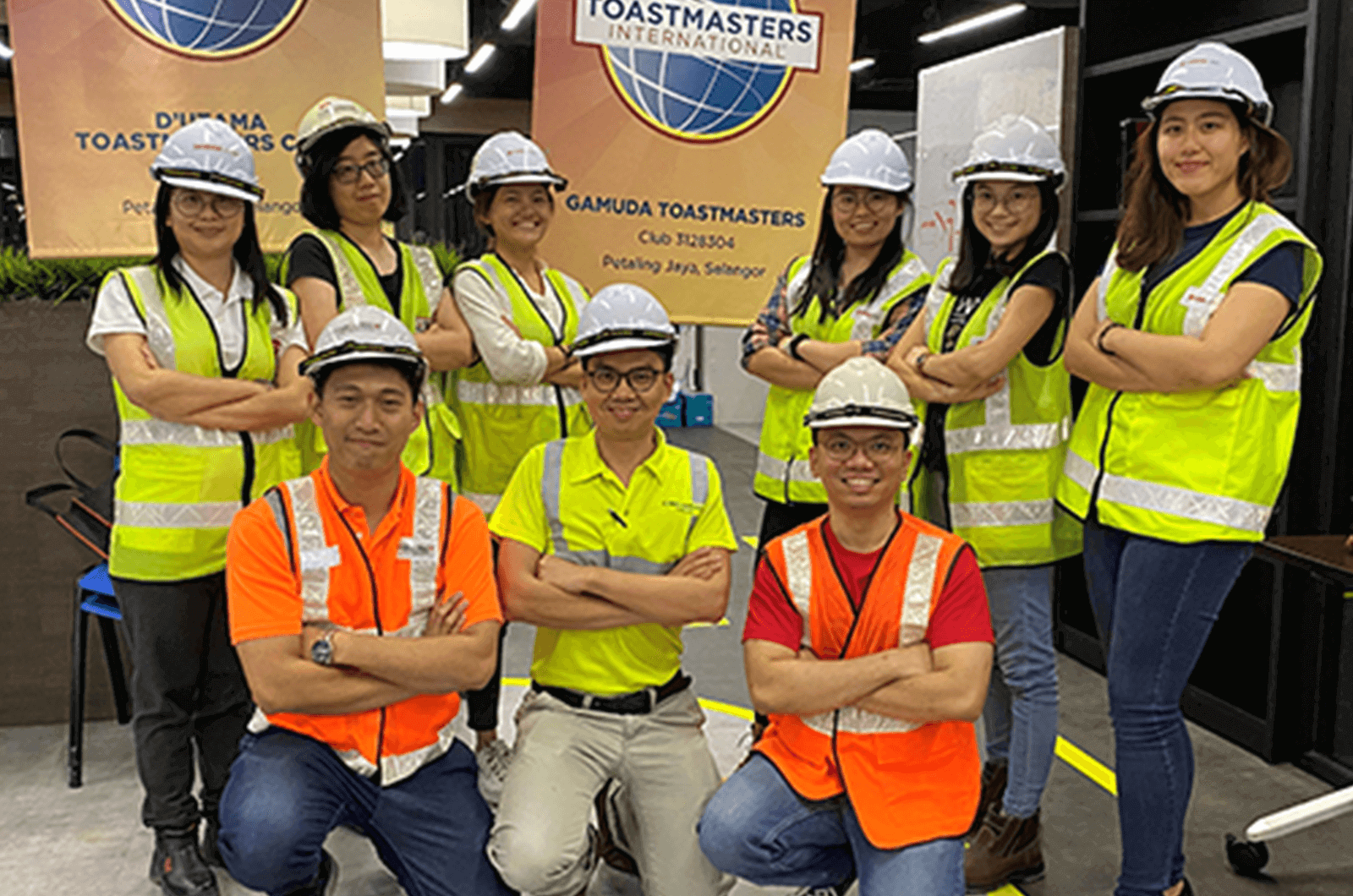 Group of Toastmasters wearing hard hats and yellow and orange vests pose in front of banners