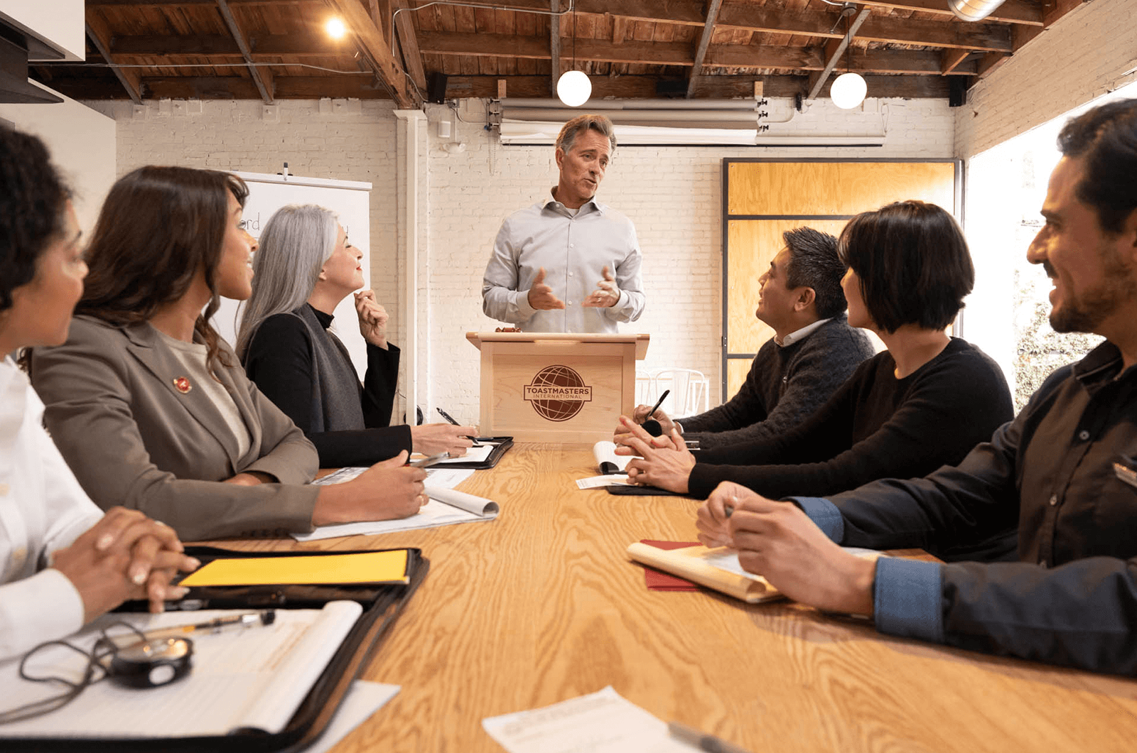 Group of people sitting around conference table while man speaks at a lectern  