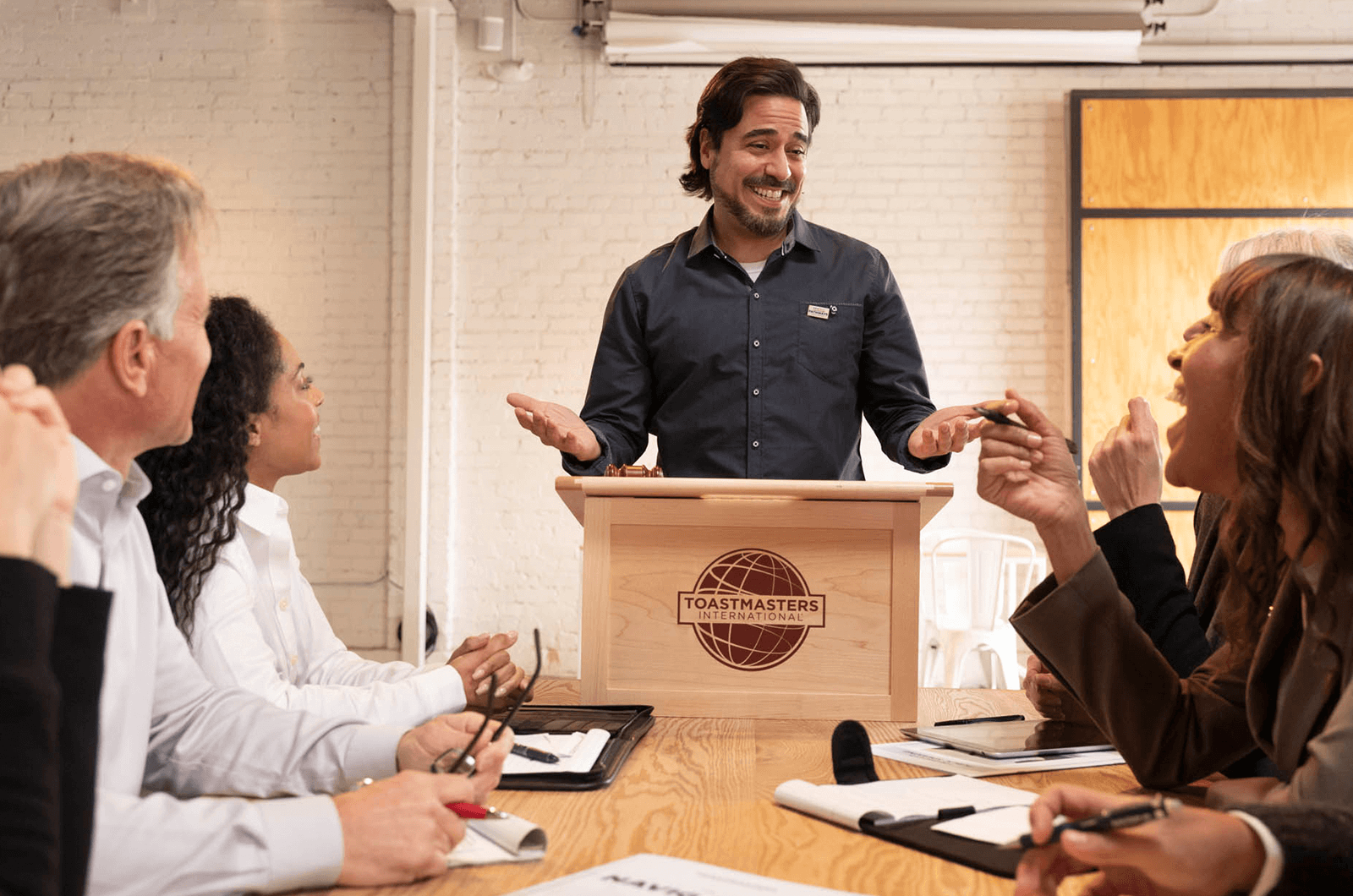 Man standing at lectern telling joke to people around table