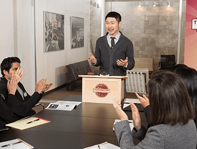 Man standing at lectern speaking to group of people clapping