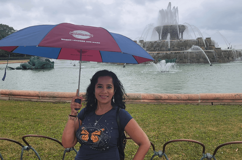 Deepa Venkat, DTM, of Frisco, Texas, shows her Toastmasters pride while staying dry at the Buckingham Fountain in the center of Grant Park in Chicago, Illinois.